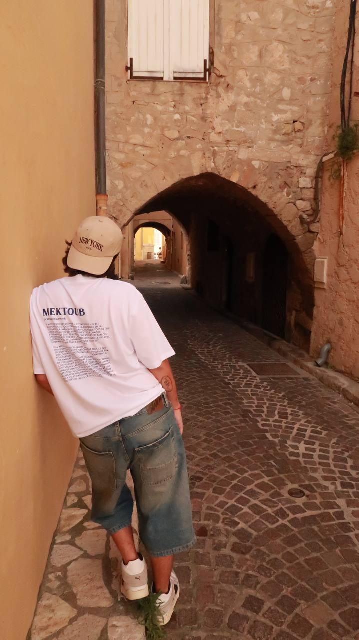 T-shirt streetwear unisexe blanc à texte imprimé, porté dans une ruelle pavée d'Antibes, style urbain et artisanal.